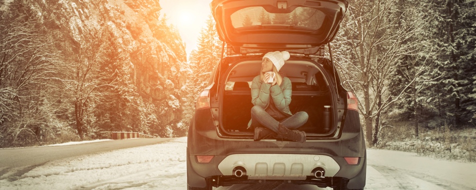 A woman in a winter hat and jacket enjoys a warm drink from a mug while sitting in the open hatchback of an SUV