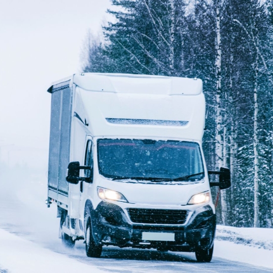 A white commercial truck drives towards the camera on a snow-covered road during a winter storm